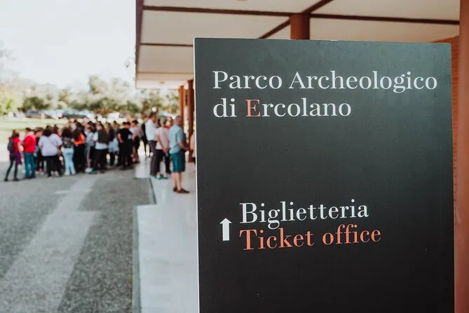 A sign reading Parco Archeologico di Ercolano, Biglietteria, Ticket office, with a group queuing behind